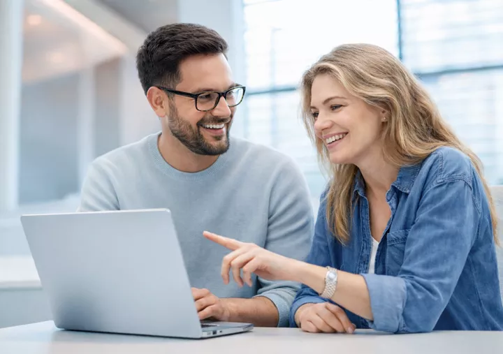 Two colleagues sit together at a desk, smiling as they look at a laptop in a bright office setting.
